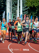 Zdjęcie, stadion, na którym biegnie grupa sportowców, za nimi flagi i drzewa.