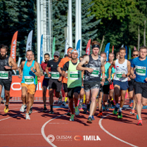 Zdjęcie, stadion, na którym biegnie grupa sportowców, za nimi flagi i drzewa.