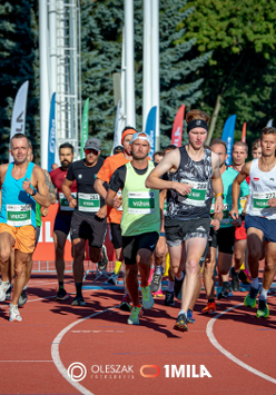 Zdjęcie, stadion, na którym biegnie grupa sportowców, za nimi flagi i drzewa.