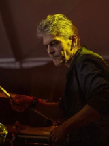 Mężczyzna w średnim wieku, z siwymi włosami i kolczykami w uchu. Ubrany w czarną koszulę z podwiniętymi rękawami. Mężczyzna gra na perkusji.