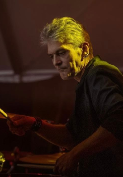 Mężczyzna w średnim wieku, z siwymi włosami i kolczykami w uchu. Ubrany w czarną koszulę z podwiniętymi rękawami. Mężczyzna gra na perkusji.