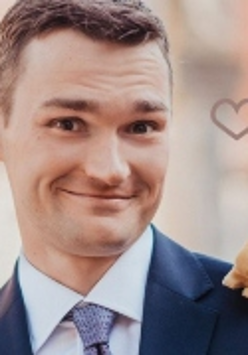 Zdjęcie przedstawia uśmiechniętego mężczyznę w marynarce i koszuli z krawatem, mającego na lewym ramieniu maskotkę wiewiórki.