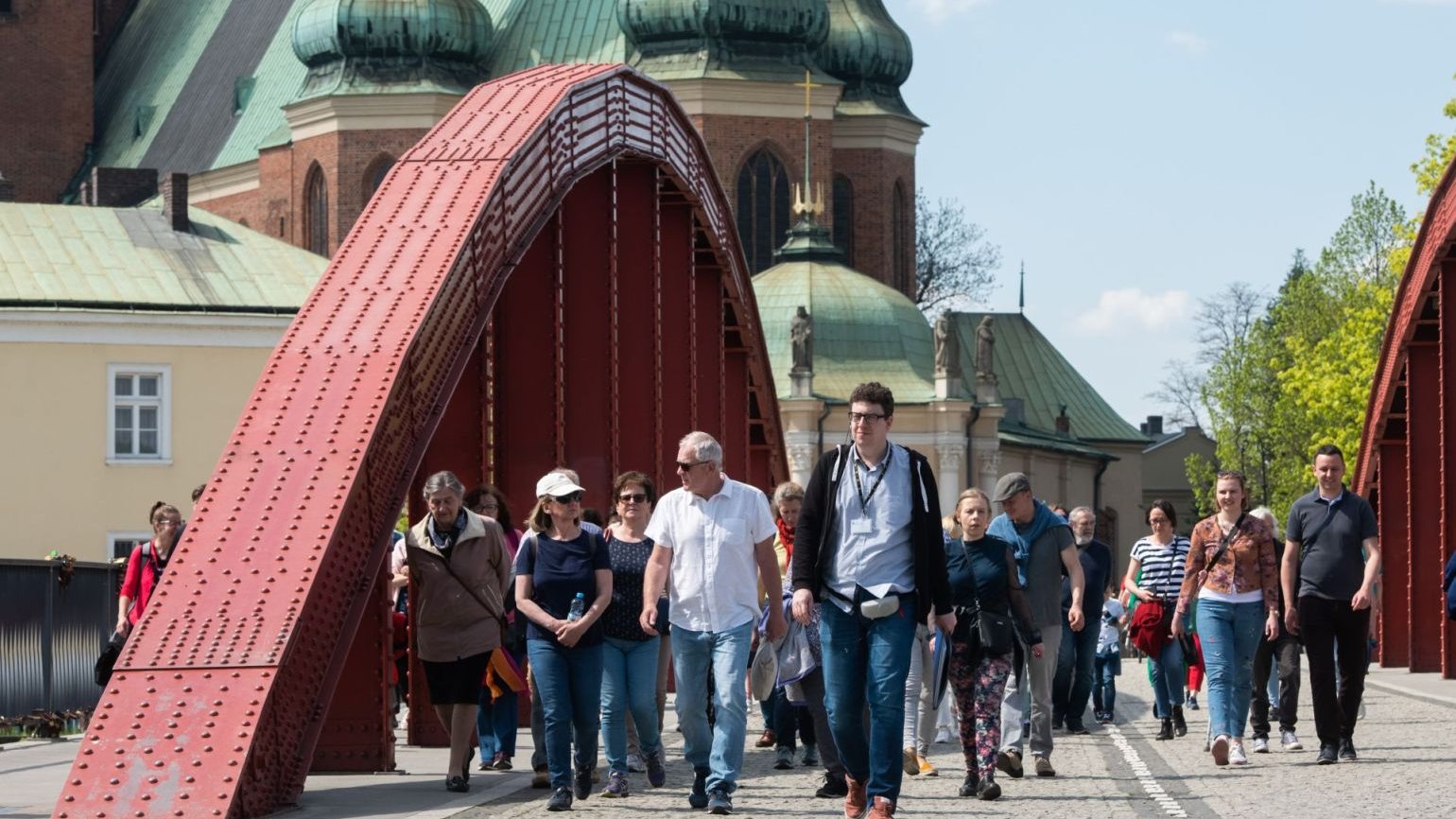 Przewodnik prowadzi dużą grupę zwiedzających przez most, w tle Katedra Poznańska