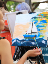 Zdjęcie, kobieta stoi przed malowanym przez siebie obrazem, na obrazie żagłówka.