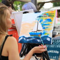 Zdjęcie, kobieta stoi przed malowanym przez siebie obrazem, na obrazie żagłówka.