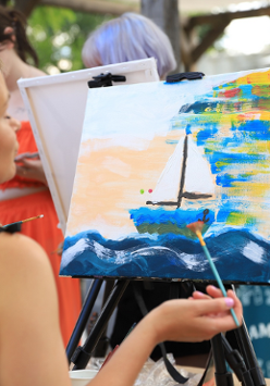 Zdjęcie, kobieta stoi przed malowanym przez siebie obrazem, na obrazie żagłówka.