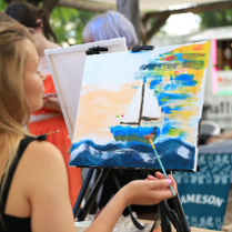 Kobieta zwrócona przodem do płótna. Maluje na nim obraz. który przedstawia łódkę na wodzie.