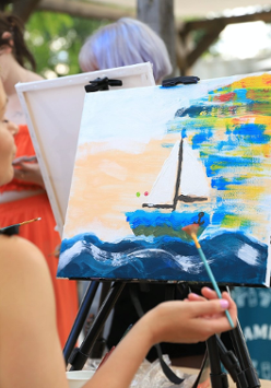 Kobieta zwrócona przodem do płótna. Maluje na nim obraz. który przedstawia łódkę na wodzie.