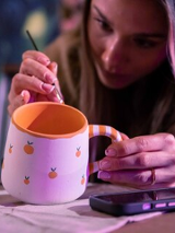Kobieta koncentrującą się na ręcznym malowaniu ceramicznego kubka. Kubek ma biały kolor z pomarańczowym wnętrzem, a jego uchwyt jest ozdobiony paskami. Dodatkowo na powierzchni naczynia znajdują się małe, pomarańczowe owoce przypominające mandarynki.
