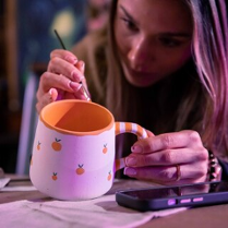 Kobieta w skupieniu maluje ceramiczny kubek cienkim pędzlem. Kubek jest biały, ozdobiony wzorem w małe pomarańcze, ma pomarańczowe wnętrze oraz ucho w biało-pomarańczowe paski. Kobieta trzyma naczynie lewą ręką, a obok niej na stole leży smartfon.
