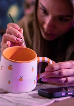 Kobieta w skupieniu maluje ceramiczny kubek cienkim pędzlem. Kubek jest biały, ozdobiony wzorem w małe pomarańcze, ma pomarańczowe wnętrze oraz ucho w biało-pomarańczowe paski. Kobieta trzyma naczynie lewą ręką, a obok niej na stole leży smartfon.