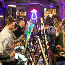 Grupa osób maluje obrazy, siedząc w pomieszczeniu o ceglanych ścianach.