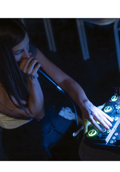 Na zdjęciu młoda dziewczyna siedzi przy konsoli didżejskiej, w ręce trzyma mikorofon, coś mowi.