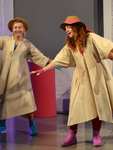 Na scenie dwie uśmiechnięte aktorki tańczą w pelerynach przeciwdeszczowych, kolorowych kaloszach i kapeluszach na głowie