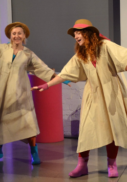 Na scenie dwie uśmiechnięte aktorki tańczą w pelerynach przeciwdeszczowych, kolorowych kaloszach i kapeluszach na głowie