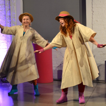 Na scenie dwie uśmiechnięte aktorki tańczą w pelerynach przeciwdeszczowych, kolorowych kaloszach i kapeluszach na głowie
