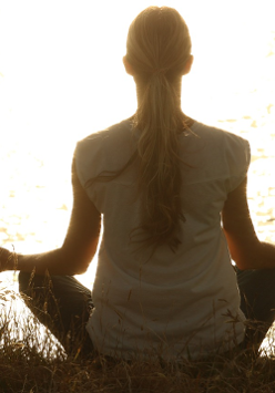 Kobieta z siedzi na brzegu jeziora i medytuje. Padają na nią promienie słońca.