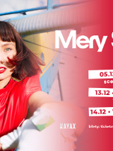 Kobieta zwrócona do obiektywu. Wyciąga dłoń. Ma krótkie brązowe włosy zakończone czerwonymi pasemkami. Ma na sobie czerwony kombinezon.