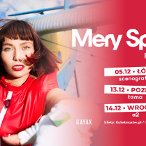 Kobieta zwrócona do obiektywu. Wyciąga dłoń. Ma krótkie brązowe włosy zakończone czerwonymi pasemkami. Ma na sobie czerwony kombinezon.