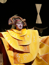 Śpiewak operowy stoi na scenie ubrany w żółty kostium. W tle kilku innych uczestników opery.