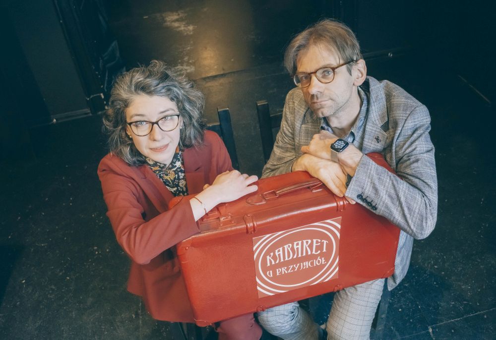 Mężczyzna i kobieta siedzą na krzesłach trzymając na kolanach czerwoną walizkę.