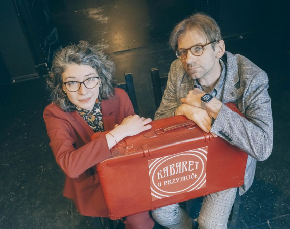 Mężczyzna i kobieta siedzą na krzesłach trzymając na kolanach czerwoną walizkę.