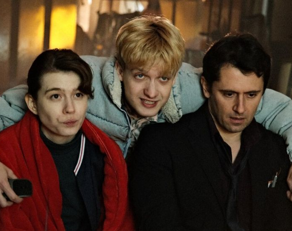 Mężczyzna pochyla się nad kobietą i mężczyzną, którzy siedzą na krzesłach. Obejmuje ich.