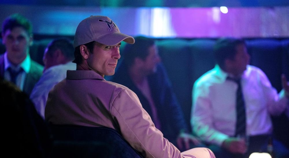 Mężczyzna w czapce z daszkiem odwraca się i spogląda na kogoś uważnie i przebiegle.