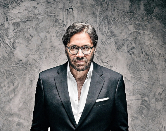 Mężczyzna w okularach, marynarce, białej koszuli, z ciemnym zarostem, patrzy w obiektyw