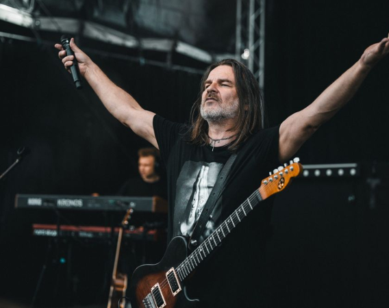 Mężczyzna z gitarą na scenie szeroko rozkłada ręce.