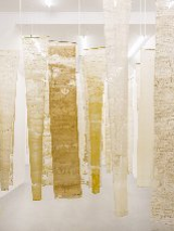 Na zdjęciu widoczna jest instalacja artystyczna składająca się z pionowo zawieszonych, półprzezroczystych tkanin w odcieniach bieli, beżu i żółci. Materiały te mają nieregularne struktury, które przypominają naturalne formy, takie jak wosk pszczeli czy pergamin. Instalacja ta buduje atmosferę kontemplacji i skupienia, symbolizując pracę pszczół i kolektywny charakter ich działań.
