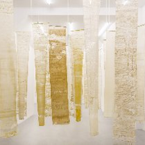 Na zdjęciu widoczna jest instalacja artystyczna składająca się z pionowo zawieszonych, półprzezroczystych tkanin w odcieniach bieli, beżu i żółci. Materiały te mają nieregularne struktury, które przypominają naturalne formy, takie jak wosk pszczeli czy pergamin. Instalacja ta buduje atmosferę kontemplacji i skupienia, symbolizując pracę pszczół i kolektywny charakter ich działań.