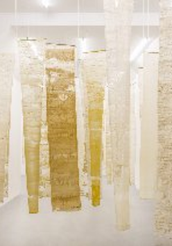 Na zdjęciu widoczna jest instalacja artystyczna składająca się z pionowo zawieszonych, półprzezroczystych tkanin w odcieniach bieli, beżu i żółci. Materiały te mają nieregularne struktury, które przypominają naturalne formy, takie jak wosk pszczeli czy pergamin. Instalacja ta buduje atmosferę kontemplacji i skupienia, symbolizując pracę pszczół i kolektywny charakter ich działań.