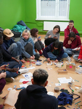 Na podłodze w pomieszczeniu siedzi grupa młodych ludzi. Wspólnie wycinają coś z papieru i rozmawiają.