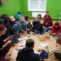 Na podłodze w pomieszczeniu siedzi grupa młodych ludzi. Wspólnie wycinają coś z papieru i rozmawiają.