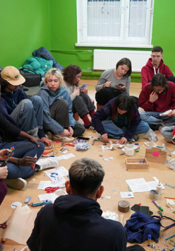 Na podłodze w pomieszczeniu siedzi grupa młodych ludzi. Wspólnie wycinają coś z papieru i rozmawiają.