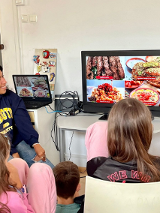 Kobieta pochodząca ze wschodu pokazuje na ekranie przysmaki ze swojego państwa. Prezentacje oglądają dzieci, które siedzą wokół na krzesłach.