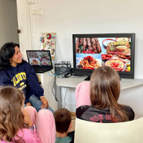 Kobieta pochodząca ze wschodu pokazuje na ekranie przysmaki ze swojego państwa. Prezentacje oglądają dzieci, które siedzą wokół na krzesłach.