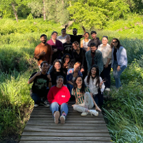 Grupa młodych ludzi stoi na drewnianym pomoście i pozuje do zdęcia. Wokół nich zielona łąka z wysokimi krzewami.
