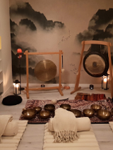 Zdjęcie kameralnej sali, w której stoją gongi i misy położone na dywanie. Na podłodze leżą białe karimaty, wałki i koce, dookoła stoją zapalone świece.