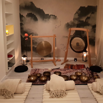 Zdjęcie kameralnej sali, w której stoją gongi i misy położone na dywanie. Na podłodze leżą białe karimaty, wałki i koce, dookoła stoją zapalone świece.