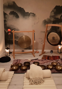 Zdjęcie kameralnej sali, w której stoją gongi i misy położone na dywanie. Na podłodze leżą białe karimaty, wałki i koce, dookoła stoją zapalone świece.