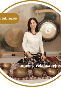 Kobieta w średnim wieku z czarnymi włosami. Ubrana w biały długi rękaw i wzorzyste spodnie. Siedzi na podłodze pokrytej dywanami. Za nią wiszą 2 gonki. Przed nią leży kilka mis.