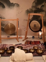Zdjęcie kameralnej sali, w której stoją gongi i misy położone na dywanie. Na podłodze leżą białe karimaty, wałki i koce, dookoła stoją zapalone świece.