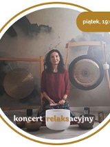 Kobieta w średnim wieku z czarnymi włosami. Ubrana w czerwony długi rękaw i wzorzyste spodnie. Siedzi na podłodze. Za nią wiszą 2 gongi. Przed nią leży kilka mis.