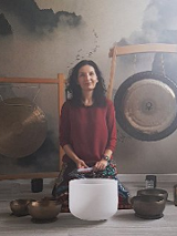 Kobieta w bordowej bluzce siedzi na podłodze. Przed nią stoją misy i gongi do grania.