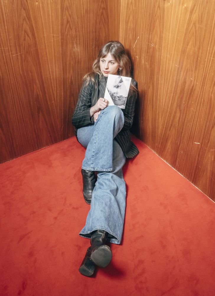 Młoda kobieta siedzi na podłodze, opiera się o ścianę. W ręku trzyma książkę.