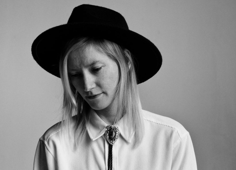 Młoda kobieta w kowbojskim kapeluszu. Dyskretnie się uśmiecha.