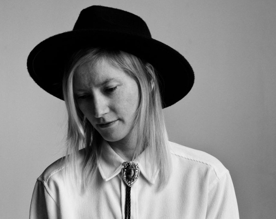 Młoda kobieta w kowbojskim kapeluszu. Dyskretnie się uśmiecha.