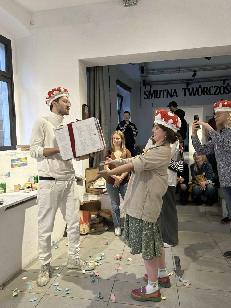 Młoda kobieta wręcza prezent młodemu mężczyźnie. Oboje mają na głowach czapki w kształcie muchomorów.